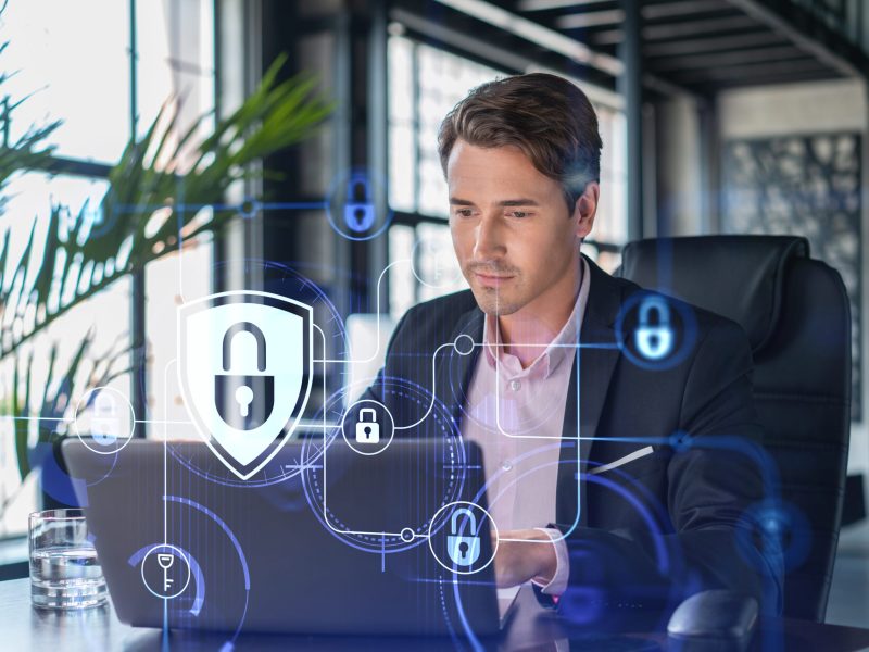 Handsome businessman in suit at workplace working with laptop to defend customer cyber security. Concept of clients information protection and brainstorm. Padlock hologram over office background.